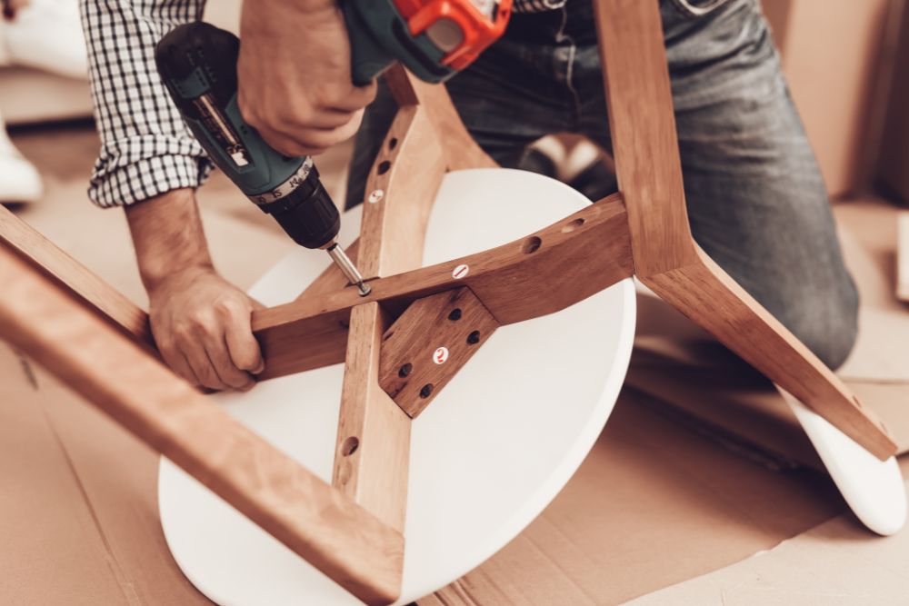 A man operates a drill while constructing a wooden chair in a workshop setting.