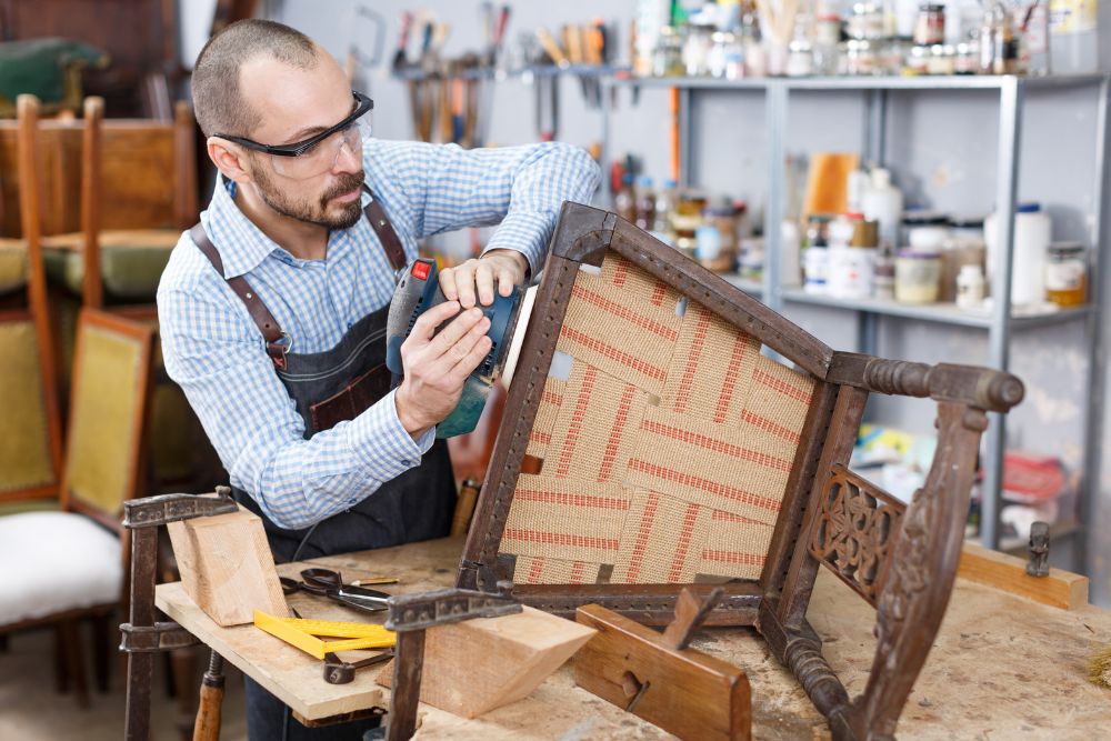 A man diligently working on a wooden chair in a workshop, surrounded by various tools and equipment.