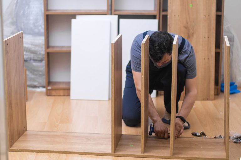A man is assembling a wooden shelf, focused on his task with tools and materials spread out around him.