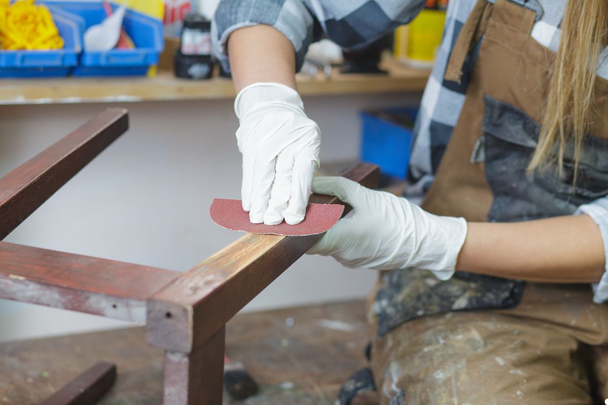 A woman wearing overalls is sanding a wooden chair, demonstrating craftsmanship in a bright workshop environment.